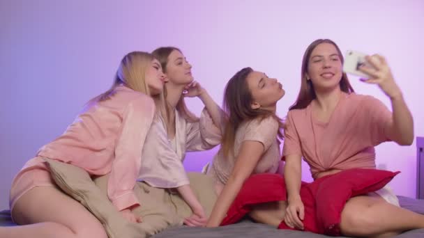 Четыре счастливые молодые девушки в пижаме сидят на кровати и делают селфи по телефону на девичнике — стоковое видео