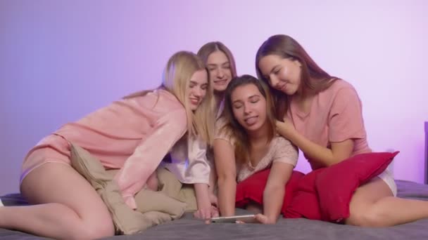 Fyra glada söta unga kvinnor i pyjamas sitter på sängen och tar selfies på telefon på möhippa — Stockvideo