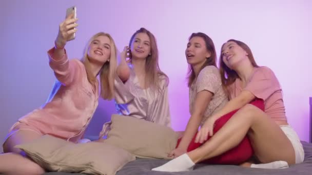 Cuatro hermosas mujeres jóvenes felices en pijama se sienta en la cama y se toma selfies en el teléfono en la despedida de soltera — Vídeo de stock