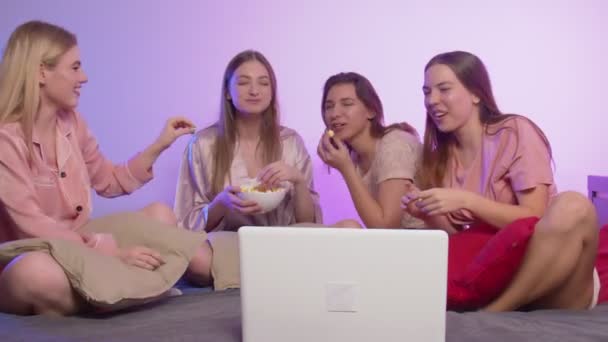 Quattro belle ragazze felici in pigiama si siedono sul letto, mangiano popcorn e guardano un film all'addio al nubilato. — Video Stock