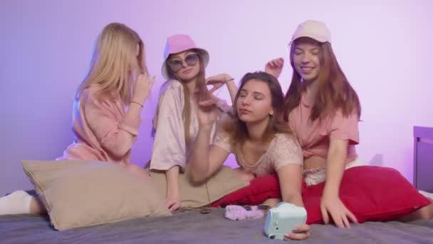 Quatro mulheres bonitas felizes no pijama senta-se na cama e toma um silfie na câmera vintage em óculos de sol e chapéus na festa de despedida de solteira — Vídeo de Stock