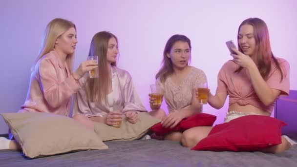 Cztery szczęśliwe, ładne, młode kobiety w piżamie siedzą w łóżku. Jedna kobieta nagrywa wideo przez telefon tosty i stuka szklankami na wieczorze panieńskim — Wideo stockowe