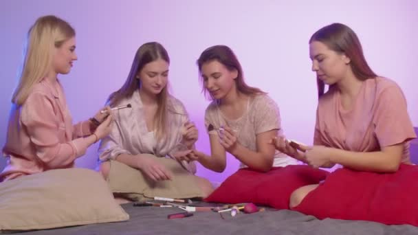 Cztery szczęśliwe, ładne, młode kobiety w piżamie siedzą w łóżku i robią makijaż na wieczorze panieńskim. — Wideo stockowe