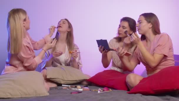 Cztery szczęśliwe, ładne, młode kobiety w piżamie siedzą w łóżku i robią makijaż na wieczorze panieńskim. — Wideo stockowe
