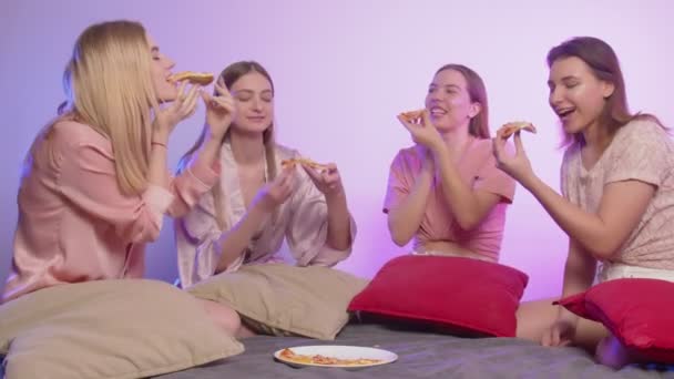 Cztery szczęśliwe, ładne, młode kobiety w piżamie siadają na łóżku, jedzą pizzę i robią zdjęcia na wieczorze panieńskim. — Wideo stockowe