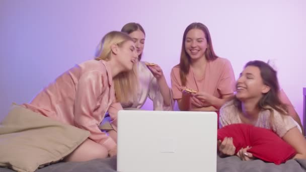 Quatro mulheres bonitas felizes no pijama senta-se na cama, assistir a um filme de comédia e come pizza na festa de despedida de solteira — Vídeo de Stock