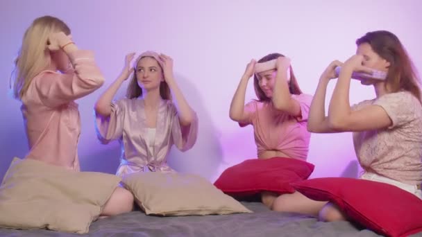Четыре счастливые молодые женщины в пижаме сидят на кровати и надевают повязки на девичник — стоковое видео