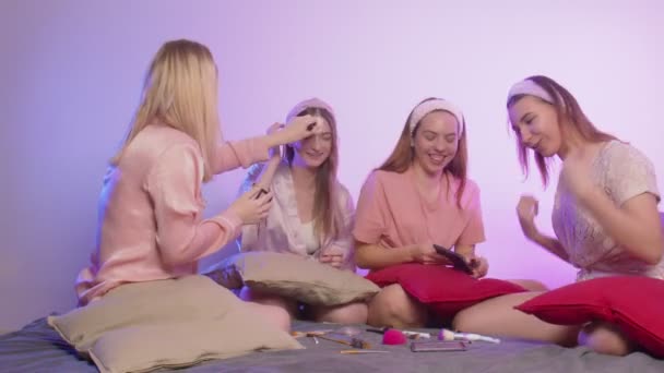 Four happy pretty young women in pajamas and headbands on head sits on bed and do make up at bachelorette party — Stock Video