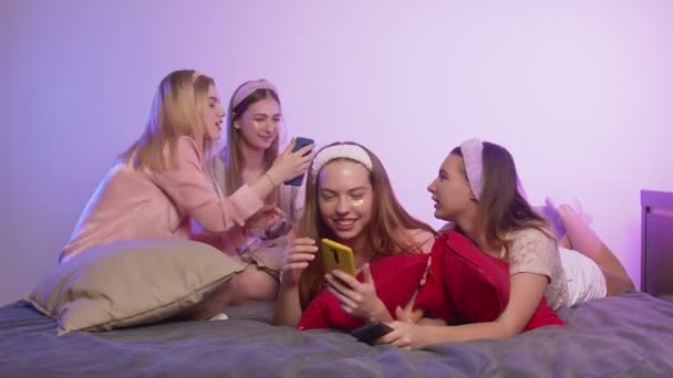 Cuatro hermosas mujeres jóvenes felices en pijama, parches de colágeno ocular y diademas en la cabeza se sienta en la cama y usa teléfonos en la despedida de soltera — Vídeo de stock