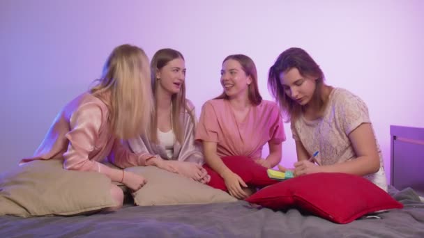 Fyra glada söta unga kvinnor i pyjamas sitter på sängen och gör en manikyr för att matcha andra på möhippa — Stockvideo