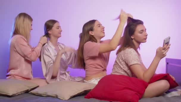 Cuatro mujeres jóvenes hermosas felices en pijama se sienta en la cama y ata coletas entre sí en la despedida de soltera — Vídeos de Stock