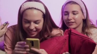 Pijamalı dört mutlu genç kadın, Göz Kolajeni bantları ve saç bantları yatakta oturuyor ve bekarlığa veda partisinde telefon kullanıyor.