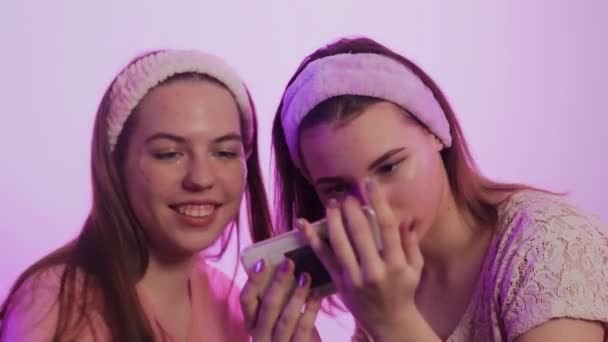 Two happy pretty young women in pajamas, Eye Collagen Patches and headbands on head sits on bed and takes photo on phone at bachelorette party — Stock Video