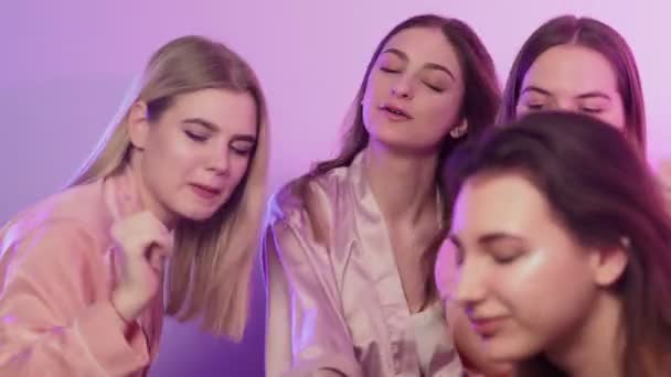 Cuatro hermosas mujeres jóvenes felices en pijama se sienta en la cama, canta, baila y graba vídeo en sus teléfonos en la despedida de soltera — Vídeo de stock