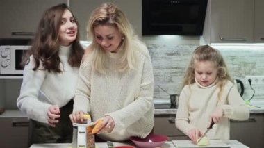 Genç anne ve iki kızı mutfakta salata pişiriyor. Anne büyük kızının havuç rendelemesine yardım eder.