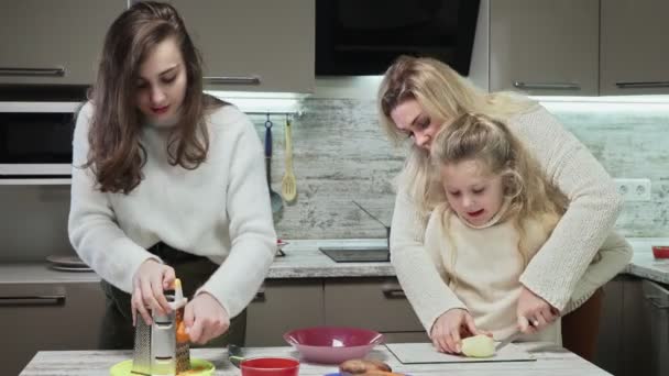 La madre giovane e due le sue figlie cucinano l'insalata a cucina. Madre aiuta la sua piccola figlia a tagliare la patata mentre la sua figlia maggiore grattugiato carota — Video Stock