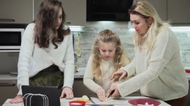 Genç anne ve iki kızı mutfakta salata pişiriyor. Anne küçük kızına havuç kesmeyi öğretiyor.