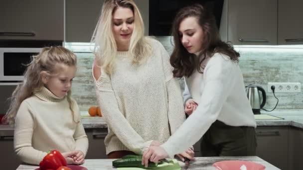 Молода мама і дві її доньки готують салат на кухні. Мати ріже огірки, розмовляючи зі своїми дочками — стокове відео