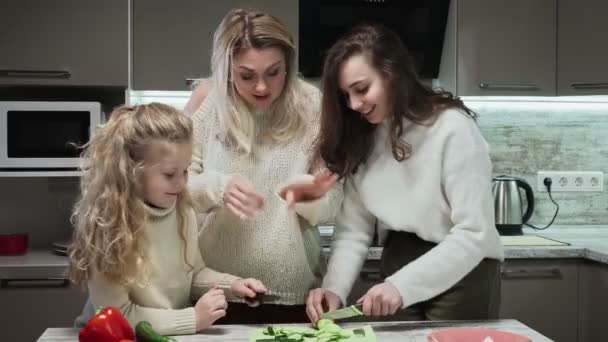 Joven madre y dos de sus hijas cocina ensalada en la cocina. Madre y sus hijas cortan pepinos — Vídeo de stock