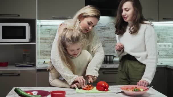 Νεαρή μητέρα και δύο κόρες της μαγειρεύουν σαλάτα στην κουζίνα. Η μητέρα μαθαίνει στην κορούλα της να κόβει κόκκινο πιπέρι. — Αρχείο Βίντεο