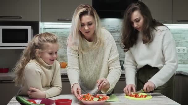 La madre giovane e due le sue figlie cucinano l'insalata a cucina. La madre mescola il salat mentre la figlia maggiore taglia un peperoncino — Video Stock