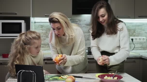 La madre giovane e due le sue figlie cucinano l'insalata di frutta a cucina. Le madri mostrano come pelare l'arancia alla sua piccola figlia — Video Stock