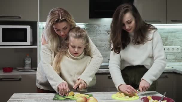 Молода мати з двома дочками готують фруктовий салат на кухні. Мама вчить свою маленьку донечку рубати — стокове відео