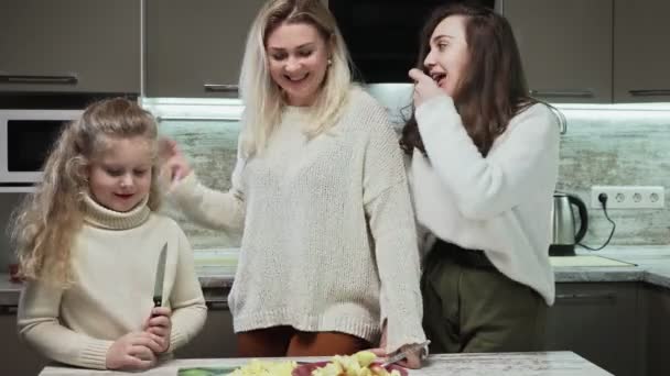 Ibu muda dan dua putrinya memasak salad buah di dapur. Gadis kecil menempatkan apel dipotong ke piring dengan salat buah — Stok Video