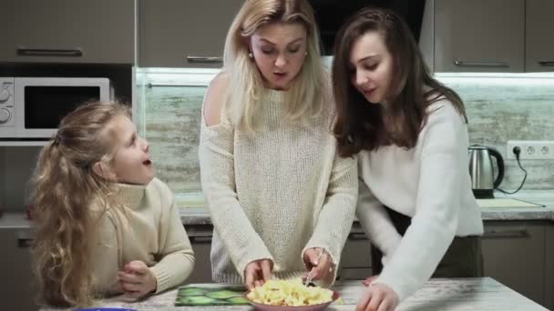 La madre giovane e due le sue figlie cucinano l'insalata di frutta a cucina. La madre mescola un salone di frutto con banane, mele e arance — Video Stock