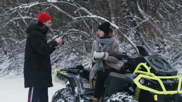 Pareja joven posando con ATV Quad bike en el bosque de invierno. Mujer bebe un té caliente — Vídeo de stock