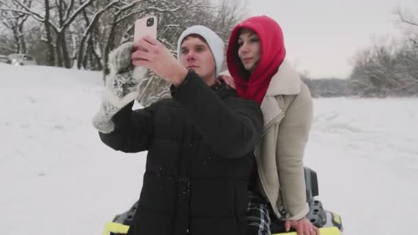 Fiatal pár pózol ATV Quad kerékpárral a téli erdőben. Egy fehér kalapos fiatalember szelfit készít a barátnőjével az ATV-n. — Stock videók
