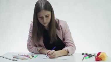 Çekici genç bir kadın beyaz bir stüdyonun masasındaki beyaz bir kağıda bir şeyler çizmek için mavi kalem kullanıyor.