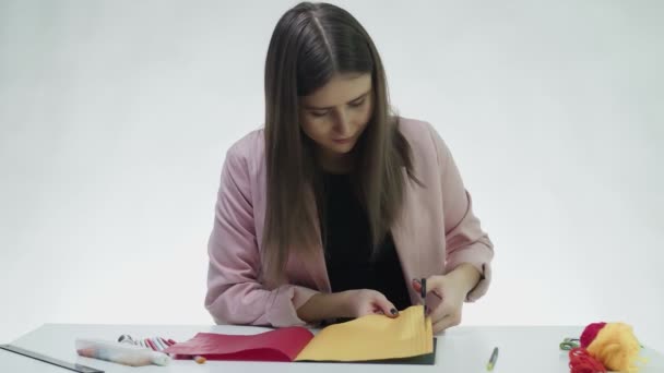魅力的な若い女性は白いスタジオでテーブルの上で色の紙の線をカットするはさみを使用しています — ストック動画