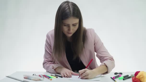 Atraktivní mladá žena používá barevné tužky a značky kreslit na bílý papír u stolu v bílém studiu — Stock video