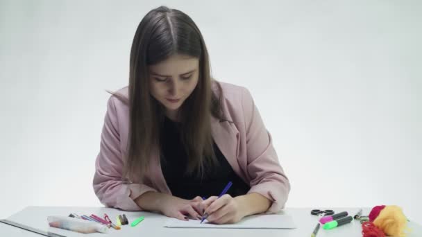 Приваблива молода жінка використовує маркери для малювання картини на білому папері за столом у білій студії — стокове відео