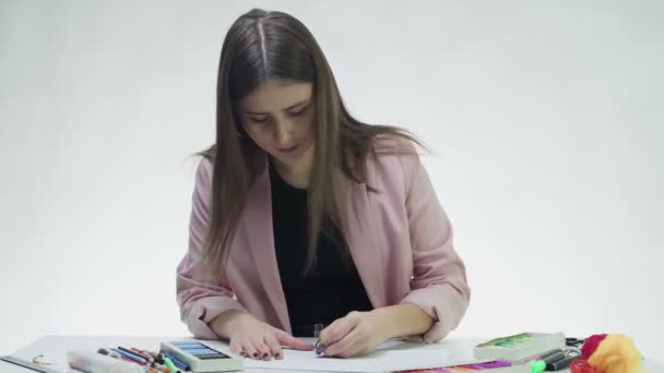 Ελκυστική νεαρή γυναίκα χρησιμοποιεί παστέλ για να ζωγραφίσει κάτι σε ένα λευκό χαρτί στο τραπέζι σε ένα λευκό στούντιο — Αρχείο Βίντεο