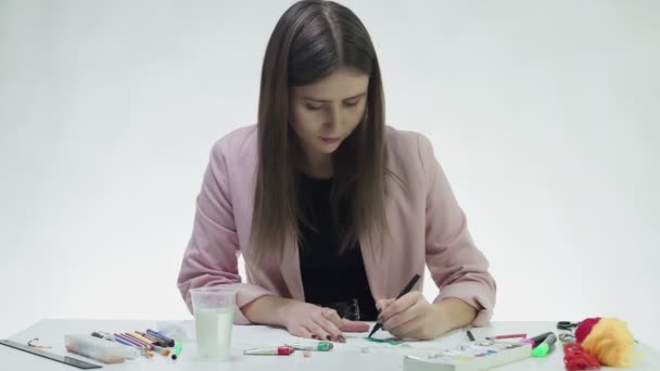 Приваблива молода жінка малює чорним маркером на білому папері за столом у білій студії — стокове відео