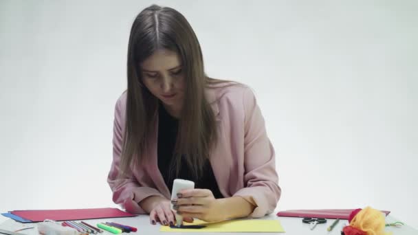 Vonzó fiatal nő csinál egy kézműves színes papír és ragasztó az asztalnál egy fehér stúdió — Stock videók