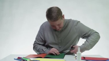 Genç adam, beyaz bir stüdyoda masada yeşil renkli bir kağıt kesmek için makas kullanıyor.