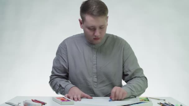 Młody mężczyzna rysuje niebieskim pastelem na białym papierze przy stole w białym studio — Wideo stockowe