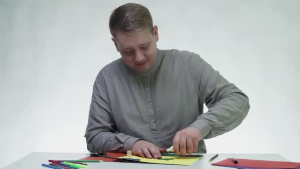 Jeune homme colle un sapin de Noël en papier coloré à une feuille jaune à la table dans un studio blanc — Video