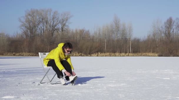 Mladá žena v slunečních brýlích a žluté bundě sedí na židli na zamrzlém jezeře a dává na bílé krasobruslení venku — Stock video