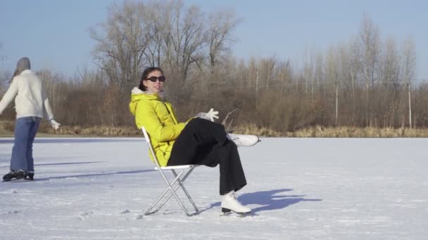 Giovane donna in occhiali da sole, giacca gialla e pattini di figura bianca si siede sulla sedia sul lago ghiacciato e indossa guanti mentre i suoi amici pattini su ghiaccio sullo sfondo — Video Stock