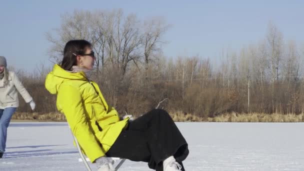 サングラス、黄色のジャケット、白いフィギュアスケートの若い女性が凍った湖の上に椅子に座ってカメラにポーズをとっている間、彼女の友人のアイススケートを背景に — ストック動画