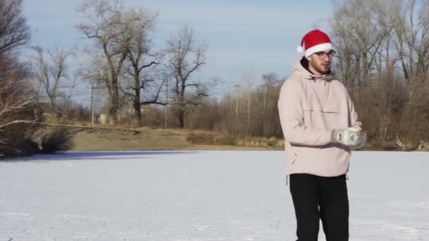 Młody człowiek w świątecznym kapeluszu jeździ na łyżwach i robi sobie selfie przez telefon na zamarzniętym jeziorze w lesie — Wideo stockowe