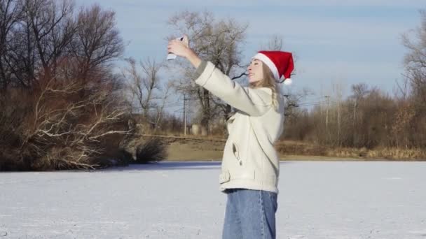 Mujer joven en Navidad santa hat paseos patinaje sobre hielo y toma selfie en el teléfono en el lago congelado en el bosque — Vídeos de Stock