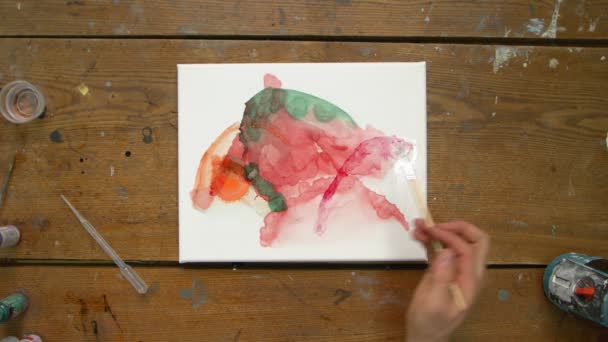 Πάνω άποψη του αρσενικού καλλιτέχνη ζωγραφίζει μια αφηρημένη εικόνα, χρησιμοποιεί πινέλο και ένα πορτοκαλί χρώμα για να ζωγραφίσει σε βρεγμένο καμβά — Αρχείο Βίντεο