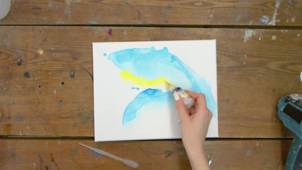 Vedere de sus a artistei de sex feminin pictează o imagine abstractă frumoasă, toarnă vopsea galbenă din tub pe pânză umedă cu forme abstracte albastre — Videoclip de stoc