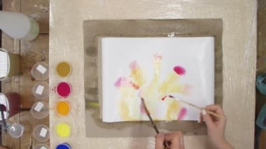 İki profesyonel ressamın üst görüntüsü Pastel kravat boyasını sarı ve kırmızı boyalarla ıslak tuvale boyuyor. Sıvı Sanat.