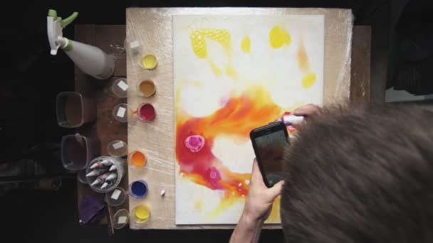 Vista superior de artista masculino pinta un cuadro abstracto, vierte un montón de pocas gotas de pintura roja a la lona húmeda y hace parte de eso en su teléfono — Vídeo de stock